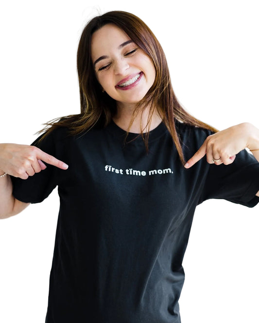 Woman wearing a black t-shirt with 'first time mom' text, pointing at it.
