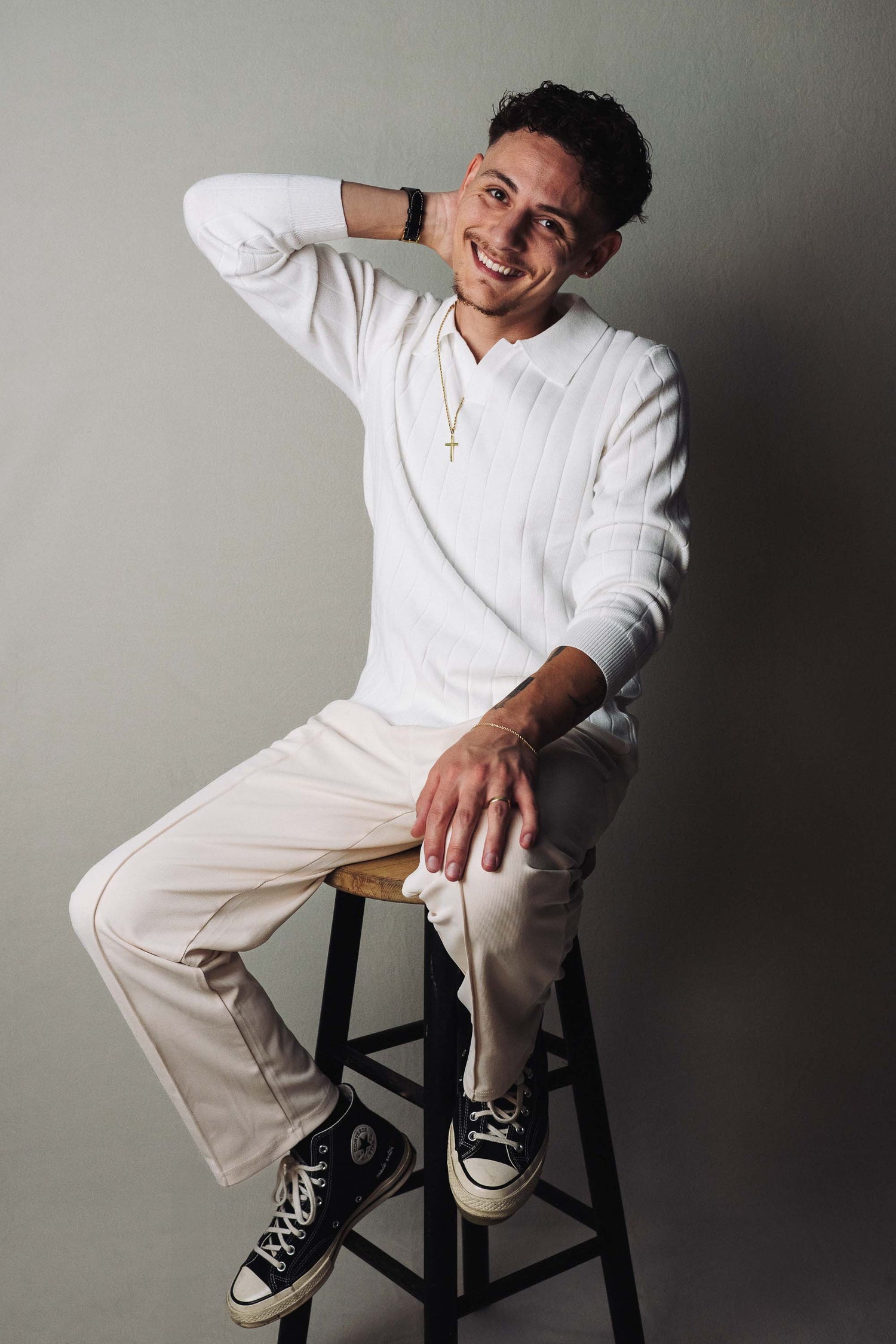 Man sitting on a stool wearing a white outfit with black sneakers against a gray background